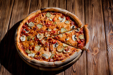 Appetizing pizza with hunting sausages, mozzarella cheese, pickled cucumbers, chili peppers on a tomato base on wood background
