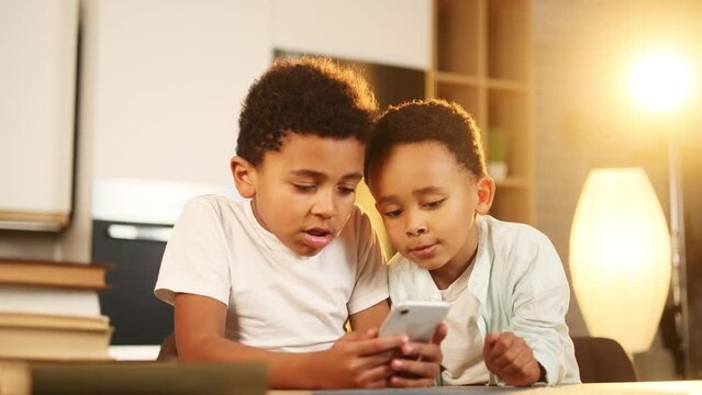 Adorable cute african american little boys brothers hold smartphone scrolling social media use educational children app application having fun with mobile phone gadget or play game together at home