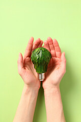 Woman's hands holding light bulb with green leaves over green background. Eco friendly lamp, energy saving, earth day concept.