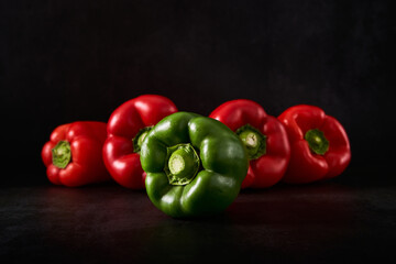 One green pepper in the foreground and four red peppers in the background. Foreground and background with black pattern. Concept for individuality or leader. Immature vs mature.