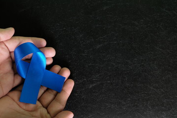 Human hands holding dark blue ribbon color on dark black background. Colon and colorectal cancer awareness and hope concept.