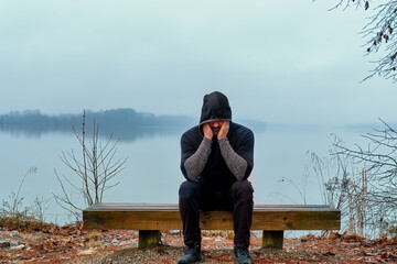 An older depressed man sitting alone on a foggy morning lake bench wearing a black hoodie.