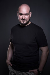 Naklejka premium Smiling bald man with a beard stands on a black background. Brutal guy in a black T-shirt. Vertical.