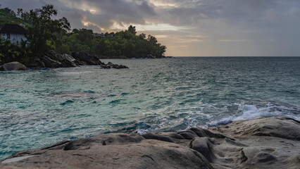 Evening seascape. Granite boulders on the shore of the turquoise ocean. A house is visible among the green vegetation. The sky is highlighted in pink, orange. Seychelles. Mahe.