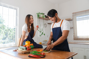 Obraz na płótnie Canvas Happy asia young couple cooking together with vegetables in cozy kitchen, preparing vegetarian food colorful variety of vegetables and herbs lying on wooden kitchen table, love and valentine concept.