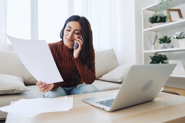A woman at home sits in front of her laptop and pays her utility bills while talking on the phone, online technology for comfort in life