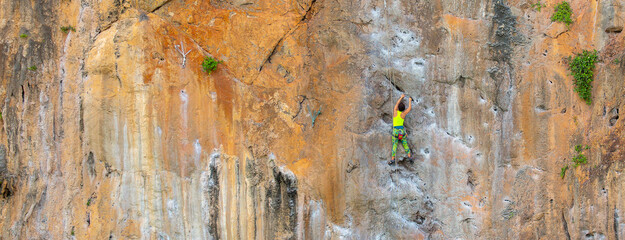 A woman climber climbs a mountain along a steep cliff. The athlete climbs a vertical stone wall with a safety rope. Extreme sport.