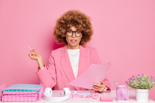 Amazed female business manager with Afro hair, makes report and holds papers, feels confused, sits at messy desktop, poses over pink background
