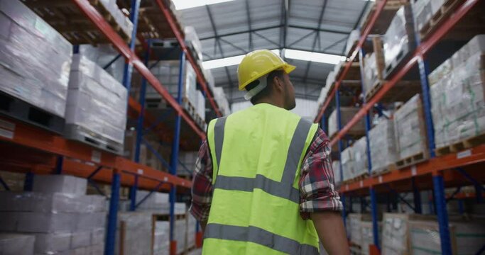 Rear view portrait of warehouse worker wearing hardhat and reflective jacket walking between storage shelves