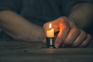 A woman's old hand holds a candle in a cartridge from a light bulb on a table at home in the dark...