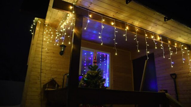Christmas tree stands by entrance of wooden residential house decorated with Christmas lights garlands at night. Real time video. Selective focus.  Winter holidays building decoration theme.