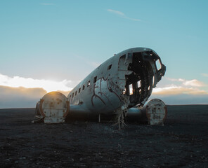 In this picture you can see an old, broken, emergency loaded airplane. Many parts are missing from the plane. The plane lies on a large black sand beach. The main colors are gray and blue.