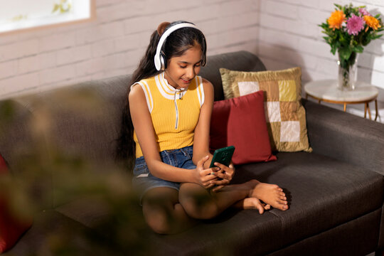 Portrait Of A Happy Girl Watching Video On Mobile Phone While Sitting On Sofa In Living Room