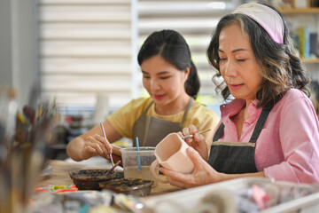 Senior artisan with Young Asian woman painting ceramic mug for sale in handmade store.