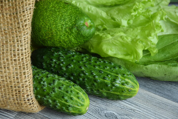 Closeup green vegetables fruit in cotton linen fabric on wooden table indoors. Pile cucumbers, avocado and bunch of lettuce in eco reusable bag day light. Ingredients for green salad. Healthy eating