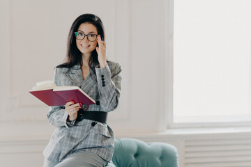 Smiling female business reporter in formal clothes holds opened notebook and pen, writes down information or checklist, wears formal clothes, works remotely from home, prepares for formal meeting
