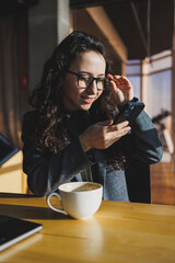 A cute woman works on a laptop in a cafe. Young concentrated brunette woman in glasses sitting at the table near the window drinking coffee. Freelance and remote work. Modern women's lifestyle