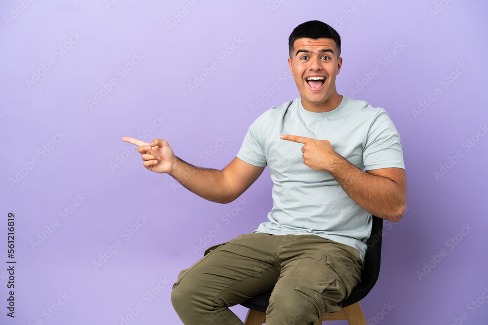 Wall mural young man sitting on a chair over isolated background surprised and pointing side