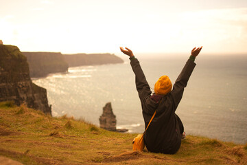 The backpacker girl with her arms raised and orange woollen hat watches the sunset. Emotions and sensations of life. Irish cliffs hit by the ocean.