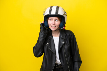 Young English woman with a motorcycle helmet isolated on yellow background listening to something by putting hand on the ear