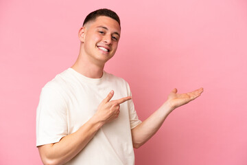Young Brazilian man isolated on pink background holding copyspace imaginary on the palm to insert...