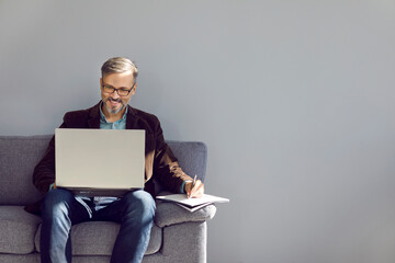 Mature businessman using laptop computer. Happy man in glasses sitting on sofa near grey copy space wall, working on modern notebook PC, studying new information, writing it down and smiling