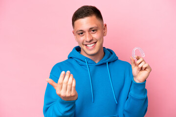 Young Brazilian man holding invisible braces isolated on pink background inviting to come with...