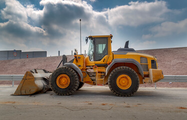 Agricultural tractor stand side on the asphalt road after working. Tractor, large yellow excavator with big wheels stand on new not yet ready road. Sunny evening with blue clouds at background