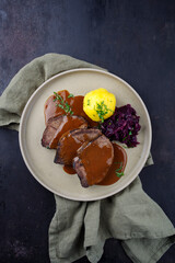 Traditionell geschmorter Sauerbraten vom Rind mit Blaukraut und Kartoffelklößen in würziger Dunkelbiersoße serviert als Draufsicht auf einem Nordic design Teller