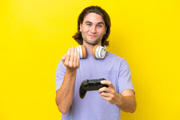 Young handsome caucasian man playing with a video game controller over isolated on yellow background inviting to come with hand. Happy that you came