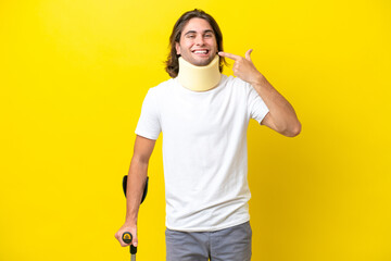 Young handsome man wearing neck brace and crutches isolated on yellow background giving a thumbs up gesture