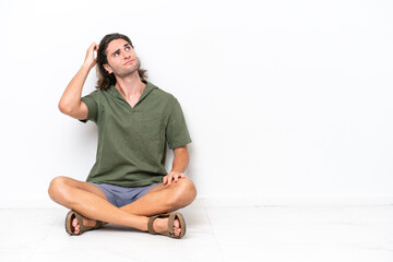 Young handsome man sitting on the floor isolated on white background having doubts and with confuse face expression