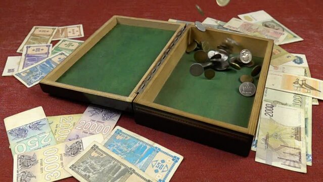Antique coins fall into a wooden box, standing on a red background among the various old banknotes, slow motion