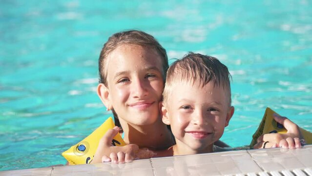 Close up of happy children swimming in pool. Brother and sister. Young beautiful girl and little boy laugh and smile broadly. Boy in inflatable armbands is holding onto side of pool. Look into camera.