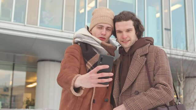 Two happy Caucasian men in love taking selfie portrait on smartphone outdoors in winter. One of men kissing his boyfriend on cheek while taking photo