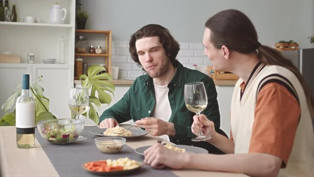 Modern Caucasian gay couple talking and drinking wine while having dinner together at home