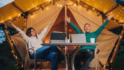 Obraz na płótnie Canvas Happy family couple freelancers working laptop on a cozy glamping tent in summer evening. Luxury camping tent for outdoor holiday and vacation. Lifestyle concept