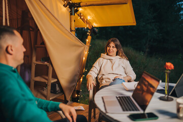Obraz na płótnie Canvas Happy family couple freelancers have a rest after working laptop on cozy glamping tent in summer evening. Luxury camping tent for outdoor holiday and vacation. Lifestyle concept