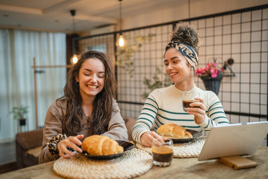 Two friends or girlfriends or sisters are eating croissant and drinking coffee for breakfast while watching something on tablet in their apartment 