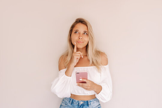 A young caucasian attractive thoughtful blonde woman holds mobile phone in her hand and thinks what to order looking up at empty copy space for text or design isolated on a beige background