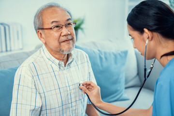 Caring young female doctor examining her contented senior patient with stethoscope in living room....