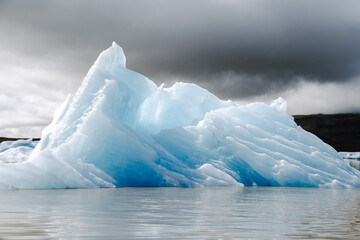 View of a blue iceberg broken off from the glacier tongue of Fjallsjökull in the glacier lagoon Fjallsárlón in Iceland on a cloudy day. With a view of Öræfajökull in the background.