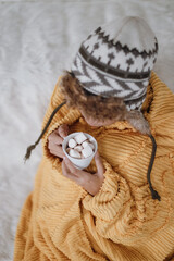 Woman wearing knitted beanie hat and using blanket holding a cup of hot chocolate with marshmallow in winter season 