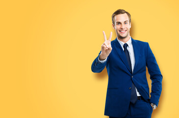 Excited businessman in confident suit showing two fingers or victory sign gesture, over yellow...