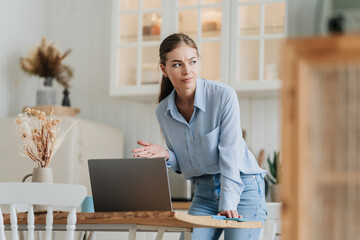 Annoyed blonde young woman in blue shirt and jeans standing at desk with laptop at kitchen with...