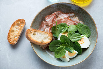 Plate with fresh spinach, bacon, hard-boiled eggs and ciabatta, elevated view on a grey granite background, horizontal shot