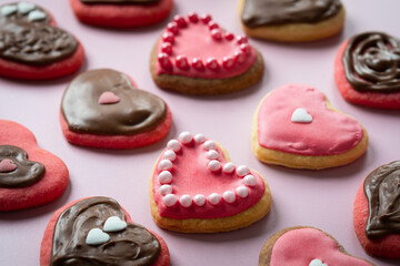 Many cookies in shape of hearts decorated by pink icing, chocolate and sprinkles on solid pale pink background