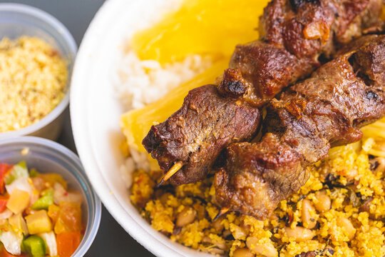 Churrasquinho brasileiro. Espetinhos com carne bovina sobre uma marmita de isopor com arroz, feij&atilde;o tropeiro, mandioca cozida e macarr&atilde;o. Comida brasileira, churrasco.