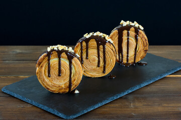 3 croissants redondos con chocolate y avellanas picadas sobre fuente de pizarra, mesa de madera y fondo negro. New York roll.
