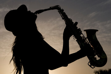 silhouette of young woman playing saxophone at sunset in backlight - obrazy, fototapety, plakaty
